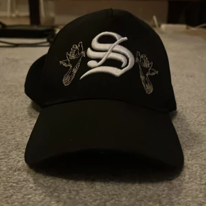 Svart keps med broderad S och fåglar - Svart keps med böjd skärm och justerbart spänne bak. Framtill finns en stor broderad vit S och två detaljerade broderade fåglar i vitt. Kepsen är tillverkad i polyester och har en stilren, urban vibe.