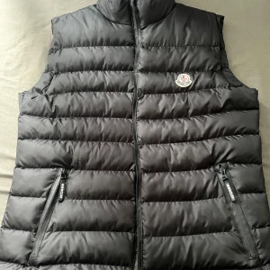 Svart dunväst från Moncler - Säljer en svart dunväst från Moncler med hög krage och dragkedja framtill. Västen har två fickor med dragkedja och Moncler-logga på bröstet. Perfekt för lager-på-lager under kyliga dagar.