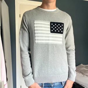 Grå stickad tröja från GANT med flagga - Snygg grå stickad tröja från GANT med amerikansk flagga i vitt och svart på bröstet. Rund halsringning och ribbade muddar. Broderad text på ena ärmen. Perfekt till jeans för en chill och stilren look. Loggo lappen i nacken sitter upp och ner men är bara fabrikat fel alltså är den äkta.  Skriv vid frågor eller funderingar🙌 fraktar inom 24h📬🤝