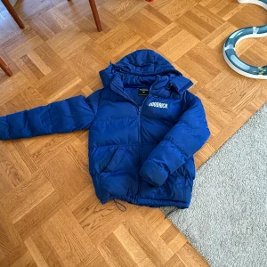 Blå pufferjacka från Hoodrich - Säljer en blå pufferjacka från Hoodrich med dragkedja och huva. Jackan har en stor logga på bröstet och ryggen. Perfekt för kyliga dagar. finns ett litet hål på innesidan bervid inefickan 