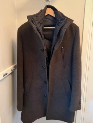 Mörkbrun rock med innerväst från Bruun & Stengade - Snygg mörkbrun rock med ståkrage och knäppning framtillsamt väst på insidan. Rocken har en innerdel med dragkedja, tre fickor framtill och en stilren, rak passform. Perfekt för dig som vill ha en klassisk look med modern twist.  Nypris är 3000kr.