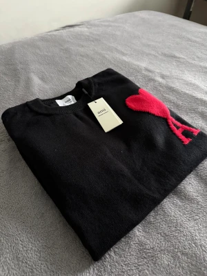 Ami Paris tröja  - Svart stickad tröja från Ami Paris  med rund hals och ribbade muddar. På bröstet finns ett stort rött hjärta med bokstaven A, vilket ger en unik och snygg detalj. Perfekt för dig som gillar stilrena plagg med en twist.