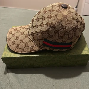 Gucci keps med GG-mönster och röd/grön detalj - KEPSEN ÄR ÄKTA! Säljer en lyxig keps från Gucci i beige och brunt med klassiskt GG-monogram över hela kepsen. Kepsen har en ikonisk röd och grön rand på sidan och justerbar rem baktill. Tillverkad i canvas med läderdetaljer. Kommer med originalbox och dustbag. 