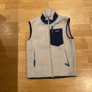 Vit fleeceväst från Patagonia XS - Säljer en vit fleeceväst från Patagonia i storlek XS. Västen har marinblå detaljer, hög krage och tre fickor med dragkedja, varav en bröstficka med logga. Materialet är mjuk fleece och västen har en clean, sportig look med blå zip-pullers.
