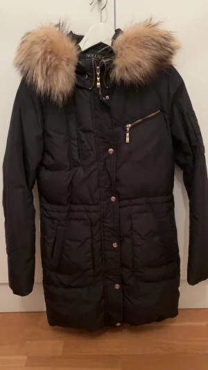 Svart dunjacka med äkta päls från Hollies - Snygg svart dunjacka från Hollies med stor fluffig äkta päls runt huvan. Jackan har guldiga detaljer, dragkedja och knappar framtill samt en bröstficka med dragkedja. Perfekt för kalla vinterdagar och riktigt stilren look. Priset kan även diskuteras!