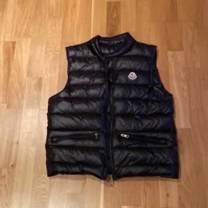 Svart dunväst från Moncler - Svart dunväst från Moncler med quiltad design och hög krage. Västen har två fickor med dragkedja framtill och Moncler-logga på bröstet. Materialet är glansigt och känns lyxigt, perfekt för att addera lite edge till din outfit.