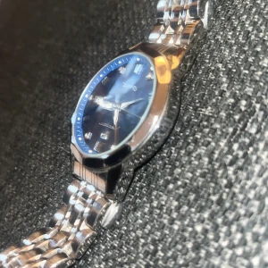 Blå och silverfärgad quartzklocka - Stilren klocka med blå urtavla och silverfärgat armband i metall. Urtavlan har romerska siffror och markerade index med stenar. Klassisk rund form och länkarmband som ger en snygg och modern look. Man får med box och duk