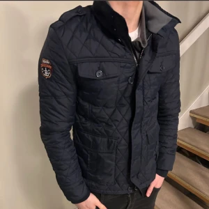Mörkblå quiltad jacka från Bendoff - Snygg mörkblå quiltad jacka från Bendoff med krage och flera fickor framtill. Jackan har ett marin-inspirerat märke på ärmen och knappar på fickorna. Perfekt för dig som gillar stilren och sportig look. S passar M