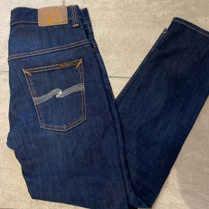 Mörkblå Nudie Jeans, storlek  - Snygga mörkblå jeans från Nudie Jeans med klassisk femficksdesign och kontrastsömmar. Bakfickorna har den ikoniska vågade brodyren i vitt. Jeansen är raka i modellen och tillverkade i slitstarkt bomullsmaterial. Perfekt för dig som gillar stilrena och tidlösa jeans. 33/30