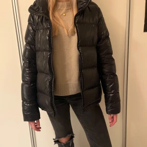 Colmar jacka Svart pufferjacka - Svart pufferjacka från Colmar med glansig finish och klassisk logga på ärmen. Jackan har dragkedja framtill, hög krage och långa ärmar. Perfekt för kalla dagar och har en snygg, lite oversized passform. Materialet är syntet och jackan är quiltad för extra värme.