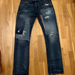 Blå jeans från Polo Ralph Lauren - Säljer ett par blå jeans från Polo Ralph Lauren med coola slitningar och patchwork-detaljer. Jeansen har en klassisk femficksdesign och en vintage look med unika revor och lagningar. Perfekta för en avslappnad stil med en twist.