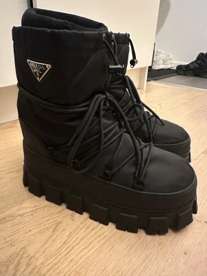 Svarta Prada moon boots med plattform - Säljer ett par svarta Prada moon boots med grov plattformssula och snörning. Skorna har quiltad ovandel med Prada-logga i metall på sidan och är fodrade för extra värme. Materialet är syntet,perfekt för kalla vinterdagar. Riktigt chunky och trendiga boots för dig som gillar statement.