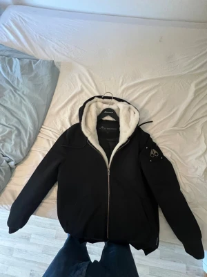 Svart teddyjacka från Moose Knuckles - Svart teddyjacka från Moose Knuckles med vit fluffig fuskpäls i huvan och insidan. Jackan har dragkedja framtill, två fickor och ett snyggt metallmärke på ärmen. Perfekt för kalla dagar med sin quiltade insida och stilrena look. Jag säljer den för den passar inte mig längre skriv ifall ni har frågor. 