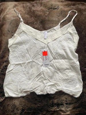 Vit linnetopp med spets  - Säljer en vit linnetopp med smala justerbara axelband och v-ringad spetskant upptill. Toppen är ärmlös och har en mjuk, lätt känsla i bomullsmaterial. Perfekt för varma dagar och enkel att styla med dina favoritjeans.