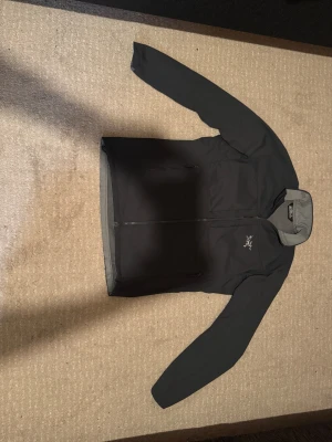 Svart vindjacka från Arc'teryx - Snygg svart vindjacka från Arc'teryx med grått foder och hög krage. Jackan har dragkedja framtill, två sidofickor med dragkedja och Arc'teryx-logga på bröstet. Perfekt för dig som gillar stilren och funktionell design.