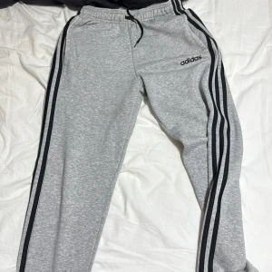 Grått sett från Adidas - Säljer en grå Adidas sweatshirt med svarta ränder längs ärmarna och stor logga framtill. Tröjan har rund hals, långa ärmar och är gjord i mjuk bomullsmix, precis som byxorna. Storlek m