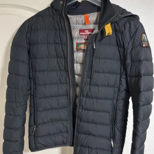 Svart lättviktsdunjacka Parajumpers  - Säljer en svart quiltad lättviktsdunjacka från Parajumpers i storlek S. Jackan har dragkedja, huva och klassisk PJS-logga på ärmen. Insidan är ljusgrå och jackan har två fickor med dragkedja. 