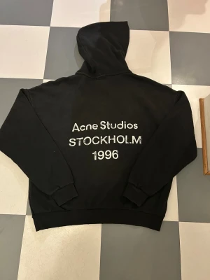 Svart hoodie Acne Studios S - Svart hoodie från Acne Studios i storlek S med vit tryckt text 'Acne Studios Stockholm 1996' på ryggen. Tröjan har huva, långa ärmar och ribbade muddar. Materialet är mjuk bomull, perfekt för chill dagar och streetwear vibes.