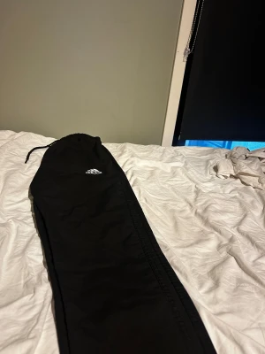 Svarta mjukisbyxor från Adidas och dragkedja  - Svarta mjukisbyxor från Adidas med vit broderad logga på vänster lår. Byxorna har elastisk midja med snörning och är gjorda i mjukt syntetmaterial. Perfekta för chill eller träning, klassisk stil med rak passform.