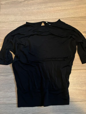 Svart blus från House of Lola - Svart blus från House of Lola i mjukt och stretchigt material. Modellen har korta ärmar och en liten öppning med knapp i nacken. Passformen är loose vid övre delen och ihopdragen runt magen, och blusen har en enkel, stilren look som funkar till många olika outfits.