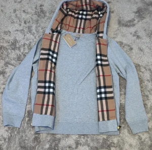 Grå Burberry hoodie med rutig huva - Snygg grå hoodie från Burberry med klassisk rutig insida på huvan i beige, svart, vit och röd. Tröjan har dragkedja, snörning vid huvan och broderad Burberry-logga på bröstet. Perfekt för dig som gillar exklusiva streetwear-vibbar.