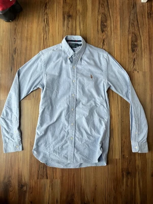 Blå skjorta från Polo Ralph Lauren - Slim fit skjorta i ljusblå bomull från Polo Ralph Lauren med klassisk krage och broderad logga på bröstet. Långärmad modell med knappar framtill och på manschetterna. Perfekt för dig som gillar stilrena och tidlösa plagg. Skjortan är mycket fint skick! 