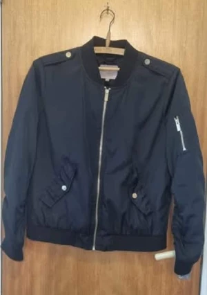 Svart bomberjacka med dragkedja - Snygg svart bomberjacka med silverfärgad dragkedja framtill och på ärmen. Jackan har ribbade muddar, två fickor med knappstängning och detaljer på axlarna. Perfekt för dig som gillar en stilren och cool look.