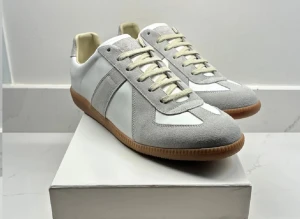 Maison Margiela  - Maison Margiela sneakers i stilren design med grå mockadetaljer och vit skinnpanel. Klassisk gummisula i brunt och snörning framtill. Snygg retrovibe och rund tå, perfekt för dig som gillar minimalistisk stil.