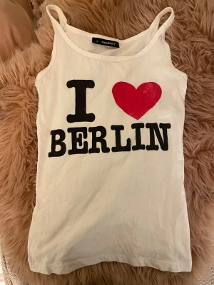 Vit linne med I ❤️ Berlin-tryck - Säljer ett vitt linne från nakedshirt i storlek S med smala axelband och stort tryck på framsidan: I ❤️ BERLIN. Linnet är gjort i mjuk bomull och har en klassisk, rak passform. Aldrig använt på grund av att den är för liten:(