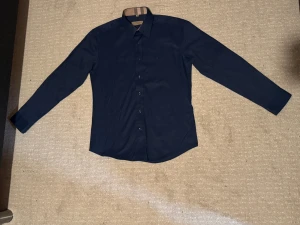 Mörkblå Burberry skjorta med rutig detalj - Snygg mörkblå skjorta från Burberry med klassiska rutiga detaljer på insidan av kragen, knappslån och ärmsluten. Skjortan har lång ärm, knappar framtill och är gjord i mjuk bomull. Perfekt för dig som gillar stilrena plagg med en twist.