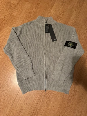 Stone Island Zip Up - Säljer en helt oanvänd grå stickad kofta från Stone Island i storlek S. Koftan har hög krage och långa ärmar, perfekt för lager på lager under kyliga dagar. Vid fler frågor är det bara att höra av sig🙌