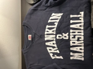 Mörkblå sweatshirt Franklin & Marshall - Snygg mörkblå sweatshirt från Franklin & Marshall med vit tryckt logga framtill. Tröjan har rund hals, ribbade muddar och långa ärmar. Perfekt för dig som gillar klassisk streetstyle och vill ha en bekväm och cool look.
