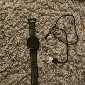 Svart smartklocka med laddkabel - Snygg svart smartklocka med fyrkantig display och silikonarmband. Klockan har magnetisk laddning och USB-kabel medföljer. Perfekt för dig som vill ha koll på träning, notiser och hälsa direkt på handleden.