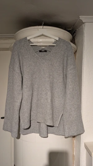 Grå stickad tröja från Bik Bok - Mysig grå stickad tröja från Bik Bok med v-ringad hals och långa ärmar. Tröjan har en loose passform och breda ribbade muddar vid ärmslut och nederkant. Perfekt för kyliga dagar och enkel att matcha med jeans eller kjol.