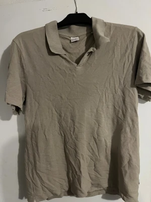 Beige henley från NN07 i bomull - Snygg beige henley från NN07 med klassisk krage och korta ärmar. Skjortan är tillverkad i mjuk bomull och har en enkel, stilren design utan mönster eller detaljer. Perfekt för dig som gillar minimalistisk stil och neutrala färger.