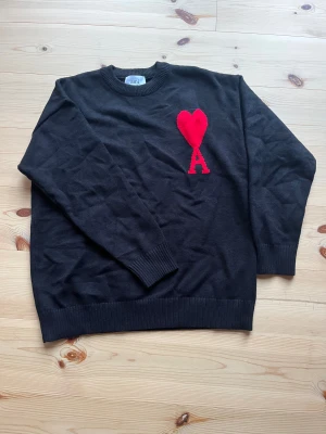 Svart stickad tröja från Ami - Svart stickad tröja från Ami med rund hals och långa ärmar. På bröstet finns ett stort rött hjärta med bokstaven A, vilket ger en unik och snygg detalj. Tröjan har ribbade muddar vid hals, ärmslut och nederkant. Perfekt för dig som gillar stilrena plagg med en twist.
