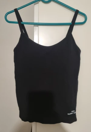 Svart linnetopp med justerbara band - Svart linnetopp med smala, justerbara axelband och enkel design. Toppen har en rak passform och är tillverkad i mjukt bomullsmaterial. Liten vit text 'every moment matters' nere vid kanten. Perfekt att styla med jeans eller kjol. Med push-up-bh.