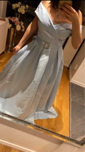 Ljusblå offshoulder långklänning - Säljer en elegant ljusblå långklänning med offshoulder och omlott upptill. Klänningen har ett brett bälte med dekorativt spänne i midjan och ett svepande, voluminöst kjolparti. Perfekt för bal eller fest! Materialet känns som satin och ger en lyxig känsla. Klänningen har även en slits och är helt ny aldrig använd, prislapp hänger kvar 
