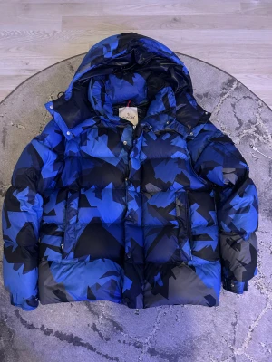 Moncler Mosa Jacka - Säljer en fet dunjacka från Moncler med blått och svart camo-mönster. Jackan har stor huva, dragkedja och tryckknappar framtill, samt Moncler-logga på ärmen. Puffig och varm design, perfekt för kalla dagar. Flera fickor och cool urban vibe.