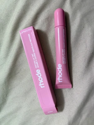 Rhode Peptide Lip Tint Ribbon - Säljer en Rhode Peptide Lip Tint i färgen Ribbon. Tuben och förpackningen är i en snygg ljusrosa nyans med vit text. Produkten är en läppbalsam med tint, perfekt för att ge läpparna en mjuk rosa ton och extra fukt. Kommer i en praktisk plasttub.