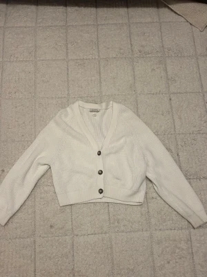 Vit kofta från Monki - Supersnygg vit kofta från Monki i stickad bomull. Koftan har v-ringning, långa ärmar och tre coola knappar framtill. Perfekt att slänga över en topp för en chill look. Passar dig som gillar enkel och stilren design.