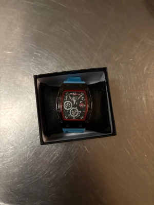 Richard Mille blå sportklocka - Säljer en Richard Mille klocka med sportig vibe och blått syntetarmband. Klockan har en rektangulär svart urtavla med röda detaljer och flera små visare. Snyggt silverfärgat spänne och cool box medföljer. Perfekt för dig som gillar statement-accessoarer.