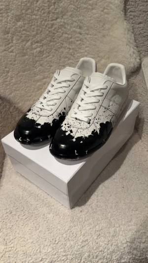 Maison Margiela svartvit sneakers - Maison Margiela sneakers i vitt skinn med svart färgstänk-design på tå och sidor. Klassisk låg modell med snörning och rund tå. Snygg kontrast mellan det vita och svarta. Box medföljs
