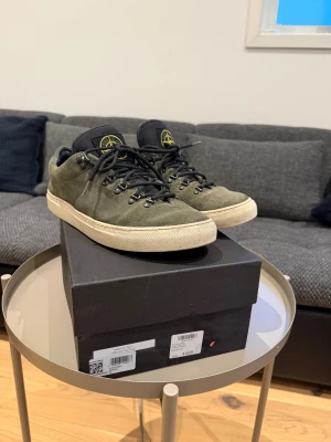 Stone Island gröna mockasneakers - Snygga gröna sneakers från Stone Island i mjuk mocka med svart snörning och robust vit sula. Tungan har en svart patch med det klassiska Stone Island-emblemet i gult. Perfekt för dig som vill ha en unik och stilren sneaker med streetkänsla.