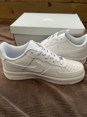 Nike Air Force 1 Low helvita sneakers - Nike Air Force 1 Low i helvit färg med klassisk rund tå och platt sula. Skorna har snörning med metallbricka märkt 'AF1', perforerad tå för ventilation och en mjuk meshfoder. Ovandel i skinn och ikonisk Nike Swoosh på sidan. Perfekt för dig som gillar clean och tidlös stil.