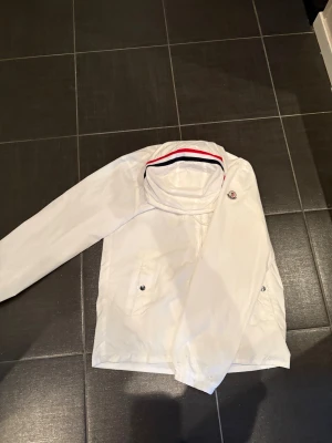 Moncler windbreaker - Letar du efter jacka till julklapp? Isåfall var AL clothing lösningen! En otroligt fet moncler jacka med de röda och marinblåa sträcken på luvan. Aldrig använd och passar perfekt nu inför jul. Jackan har några fläckar som knappt syns därav det låga priset!