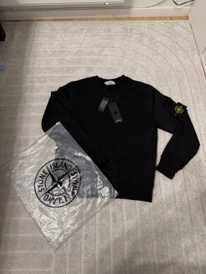Svart sweatshirt från Stone Island - Svart sweatshirt från Stone Island med klassisk rund hals och ribbade muddar. Tröjan har den ikoniska Stone Island-patchen på vänster ärm med gul och grön brodering. Tillverkad i mjuk bomull, perfekt för en clean och stilren look.
