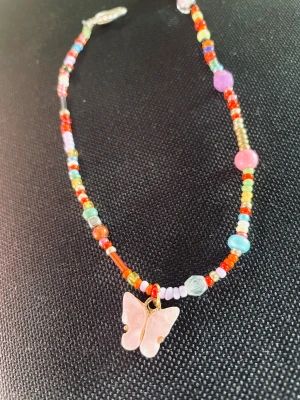 By prisförslag- slumpmässiga Färger 40 cm pärlchoker med fjäril - Unik choker halsband med små pärlor i varierande färger och former, bland annat rött, gult, lila, grönt och blått. Alla är unika så färgerna/former skiljer sig. I mitten hänger en ljusrosa fjärilsberlock med guldfärgade detaljer. Perfekt för dig som gillar lekfulla och kreativa smycken.