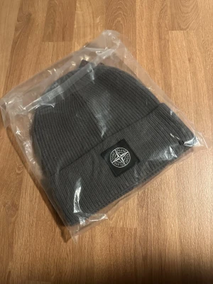 Grå ribbad mössa från Stone Island - Snygg grå mössa från Stone Island med ribbad struktur och klassisk logopatch framtill. Perfekt för dig som vill ha en stilren och enkel look med street-vibe. Materialet är mjukt och värmande, passar bra när det är kallt ute.