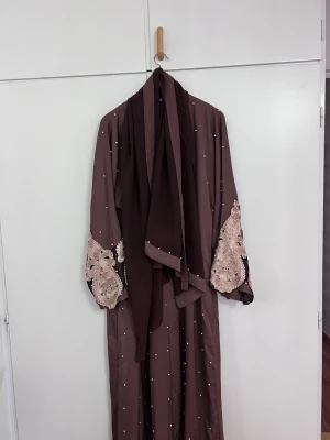 Brun långklänning med pärlor och spets - Säljer en elegant brun långklänning med pärldetaljer över hela klänningen och vackra spetsbroderier i beige på ärmar och nederkant. Klänningen har lång ärm och ett matchande sjal. Perfekt för dig som gillar unika detaljer och stilren design.